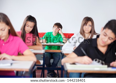 Thoughtful teenage schoolboy studying with friends in classroom