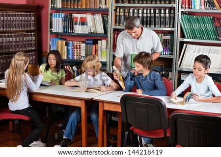 Happy mature male teacher showing book to schoolboy with students studying at table in library