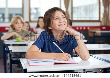 Happy young schoolboy looking up while studying at desk with friends in background