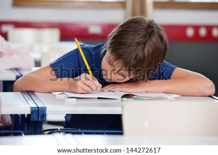 Elementary boy writing in book while leaning on desk in classroom