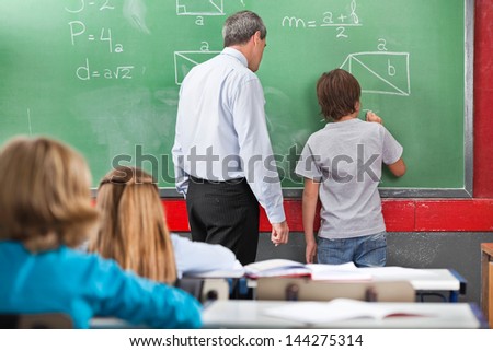 Rear view of schoolboy solving mathematics on board with teacher in classroom