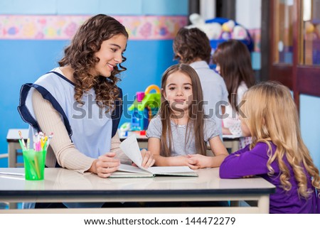 Portrait of little girl with kindergarten teacher and friend sitting at desk in classroom