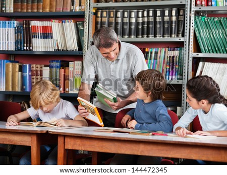Mature male teacher showing book to schoolboy with students studying at table in library