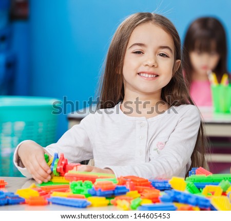 Portrait of cute little girl playing with construction blocks with friends in background at kindergarten