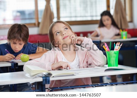 Thoughtful schoolgirl with digital tablet looking up while friends studying in background at classroom