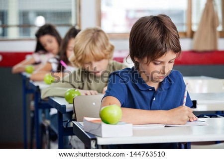 Young boy studying at desk with classmates in background at classroom
