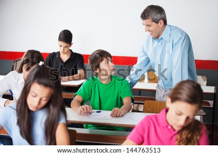 Male university student and professor looking at each other while classmates writing paper in classroom