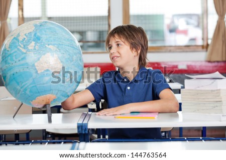 Little schoolboy searching places on globe at desk in classroom