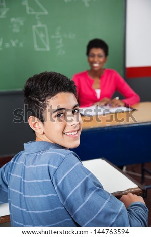 Portrait of happy teenage schoolboy sitting at desk with teacher in background