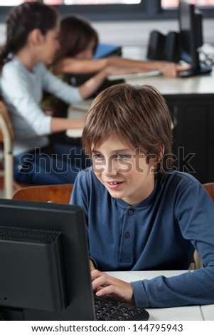 Little schoolboy using computer at desk with classmates in background