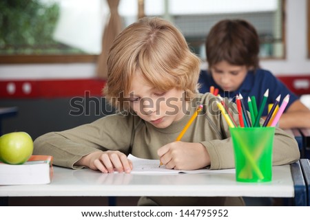 Little schoolboy writing in book at desk with classmate in background