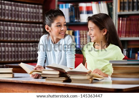 Happy cute little schoolgirls looking at each other while studying at table in library
