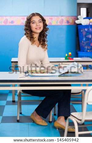 Full length portrait of young teacher with popup book sitting at desk in preschool