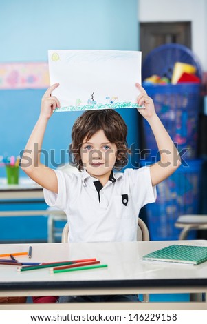 Portrait of cute boy holding drawing paper at desk in kindergarten