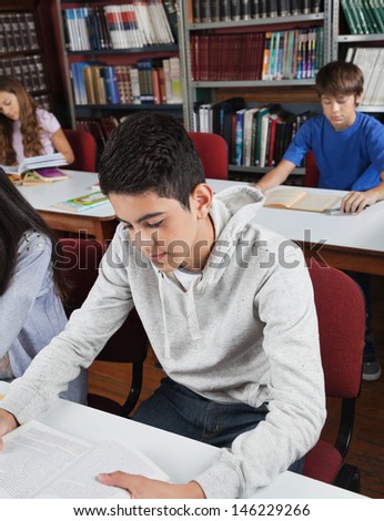 Male high school student reading book at table with classmates in library
