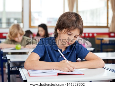 Little schoolboy writing in book at desk with classmates in background