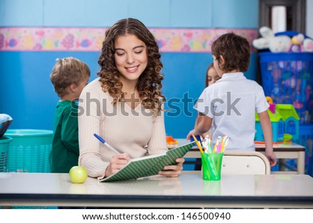 Beautiful female teacher writing in book with children in background at kindergarten