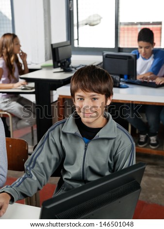 Portrait of teenage male student sitting at desk with classmates in computer class