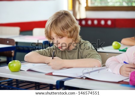 Little schoolboy writing on book with apple at desk in classroom