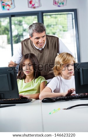 Mature male teacher looking at schoolchildren using desktop PC in computer lab