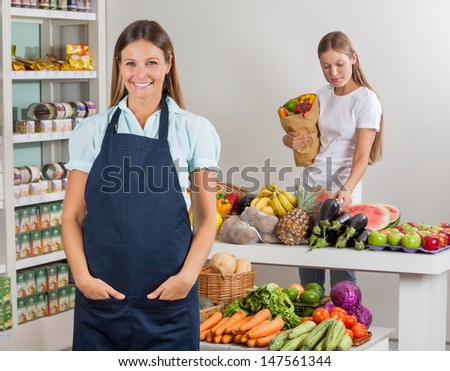 Portrait of saleswoman with hands in pocket while female customer shopping in background at supermarket