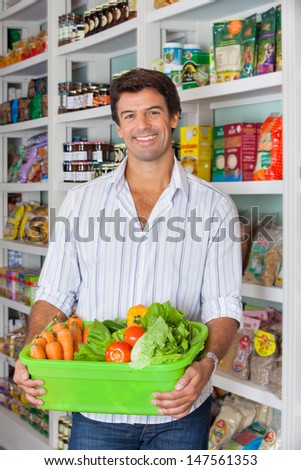 Portrait of happy mid adult male customer with vegetable basket in supermarket