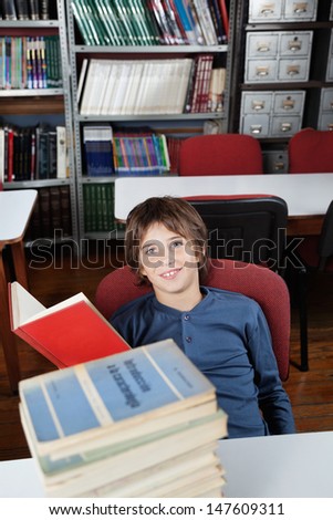 Portrait of little schoolboy sitting with stacked books at table in library