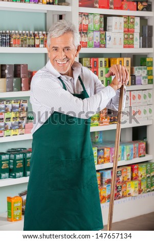 Portrait of happy senior male owner with stick standing against shelves in supermarket