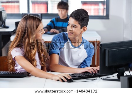 Male and female teenage friends looking at each other while using computer in classroom