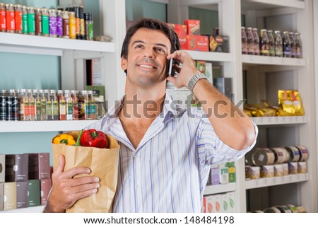 Happy mid adult man with vegetables in paper bag using cellphone in supermarket