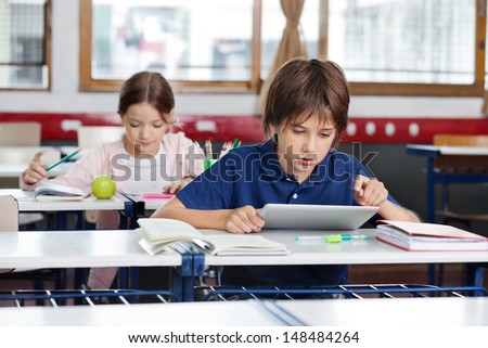 Little School Boy Using Digital Tablet With Girl Studying In Background At Classroom