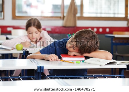Little boy sleeping on desk with girl in background at classroom