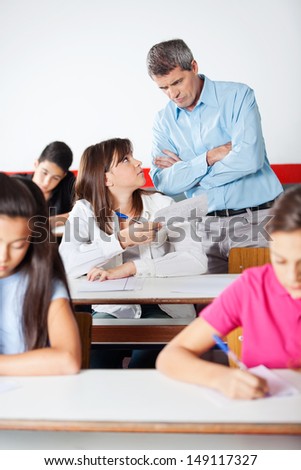 Angry mature teacher standing arms crossed while looking at student during examination in classroom