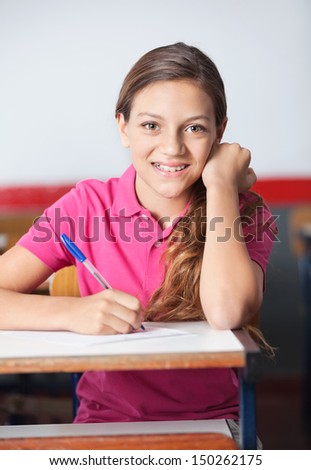 Portrait of beautiful teenage schoolgirl writing at desk in classroom
