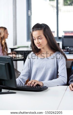 Teenage girl using desktop PC with female classmate in background at computer lab
