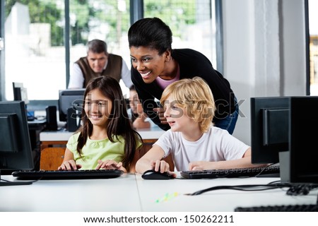 Happy Female Teacher Assisting Schoolchildren In Using Computer At School