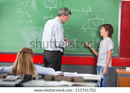 Side View Of Little Schoolboy Asking Question To Teacher While Solving Mathematics On Board In Classroom