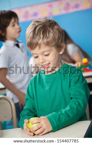 Happy cute boy holding smith apple with classmate in background at kindergarten