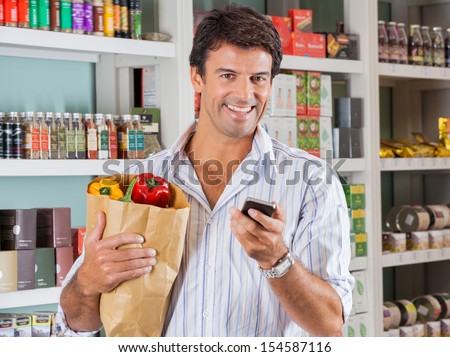 Portrait of happy male customer with mobile phone and grocery paper bag in store