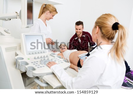 Young female doctor showing digital tablet to expectant couple with colleague using ultrasound machine in foreground
