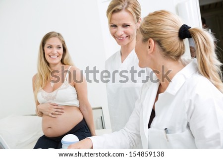 Happy doctors looking at each other with pregnant woman sitting in background at clinic