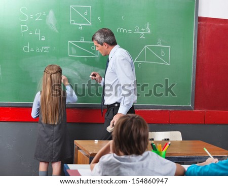 Rear of little girl solving mathematics on board with teacher looking at her