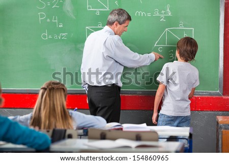 Mature male teacher assisting little schoolboy while solving mathematics on board in classroom