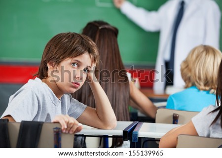 Side View Portrait Of Little Boy Leaning At Desk With Teacher And Classmates In Background