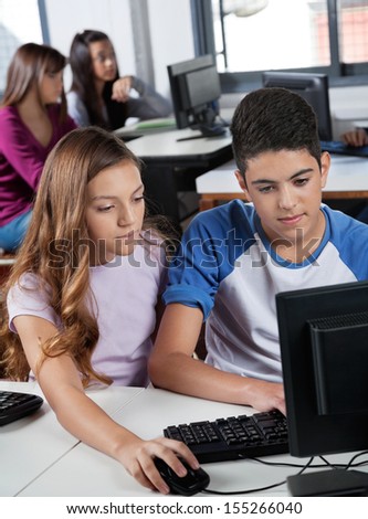 Schoolchildren using computer at desk with female classmates in background