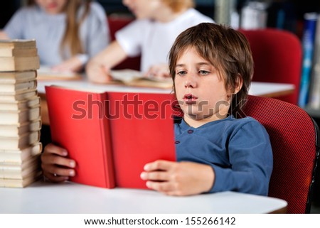 Relaxed little schoolboy reading book while sitting at table in library