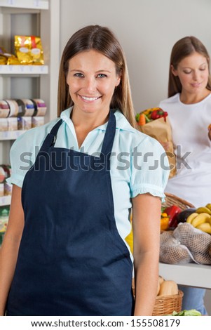 Portrait of confident saleswoman with female customer in background at supermarket