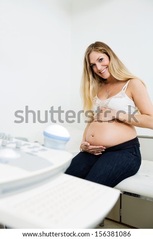 Portrait of young pregnant woman holding belly sitting by ultrasound machine in clinic