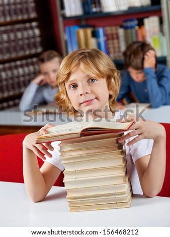 Portrait of cute little schoolboy sitting with stack of books at table in library and classmates in background