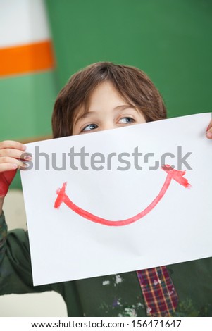 Little boy looking away while holding paper with smile drawn on it in front of face